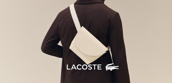 Asmuo dėvintis tamsiai rudą striukę ir per petį nešiojantis baltą krepšį ant paprasto fono.