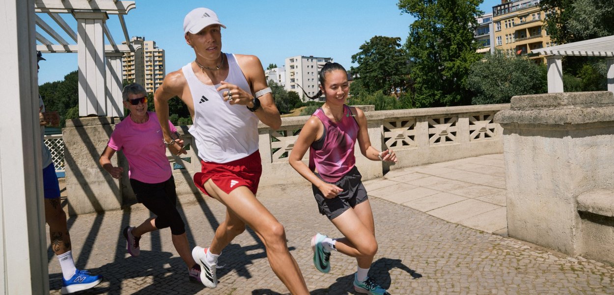 Vier Läufer in unterschiedlich sportlicher Kleidung: ein weißes Tanktop und rote Shorts, ein pinkes Oberteil, ein graues Outfit und Shorts; helles, urbanes Setting.