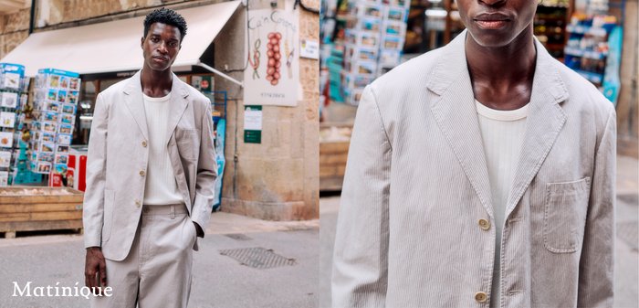 Uomo con abito in velluto a coste beige chiaro e camicia bianca in piedi su una strada con espositori di cartoline e edifici in pietra sullo sfondo.