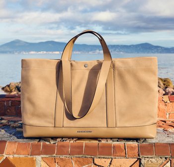 Beige Leder-Shopper mit doppelten Henkeln, der auf einer gefliesten Oberfläche am Meer liegt, im Hintergrund sind Berge sichtbar.