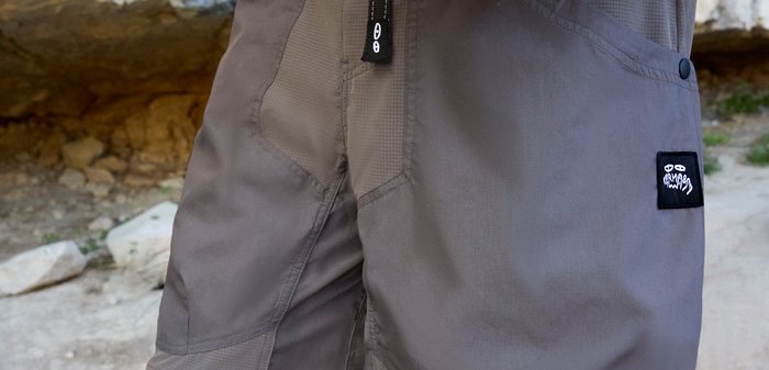 Κοντινό πλάνο γκρι παντελονιού εξωτερικού χώρου με ενισχυμένες ραφές, μπροστινό φερμουάρ και μικρό μαύρο λογότυπο στο δεξί μηρό.
