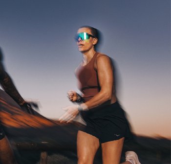 Coureur en débardeur marron et shorts noirs, portant des lunettes de soleil réfléchissantes bleues. Un flou de mouvement capture un mouvement dynamique contre un ciel dégradé.