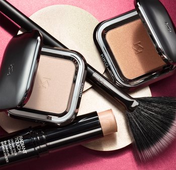 Two square compacts of face powder in silver cases, one light and one bronze. A black makeup brush and a highlighting stick are included.