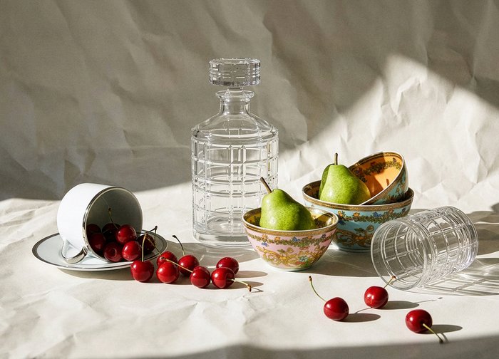 Δύο πράσινα αχλάδια σε διακοσμητικά μπωλ, χυμένες κερασιές κοντά σε ένα αναποδογυρισμένο φλιτζάνι τσαγιού και ποτήρι, και ένα διάφανο γυάλινο καράφα πάνω σε ένα ζαρωμένο ύφασμα.