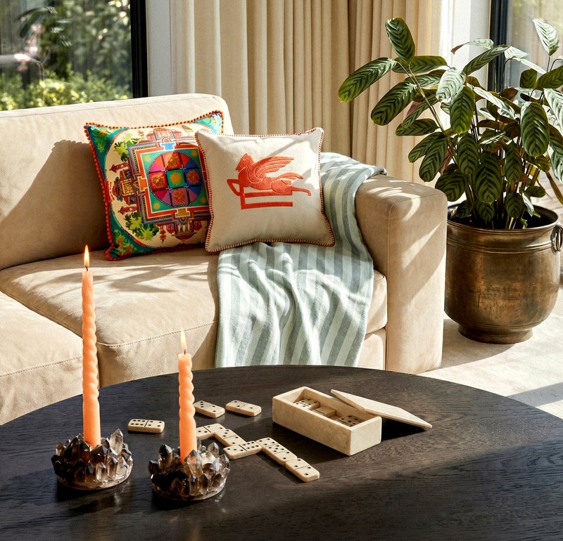 Canapé beige avec des coussins colorés et une couverture à côté d’une grande fenêtre, table ronde en bois sombre avec des bougies allumées et des dominos, et une plante en pot à proximité.