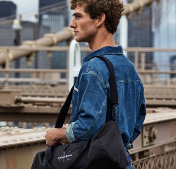 Borsa a tracolla nera con logo Calvin Klein, portata a spalla. Camicia di jeans con tonalità azzurro chiaro, tessuto strutturato, con bottoni, stile casual.