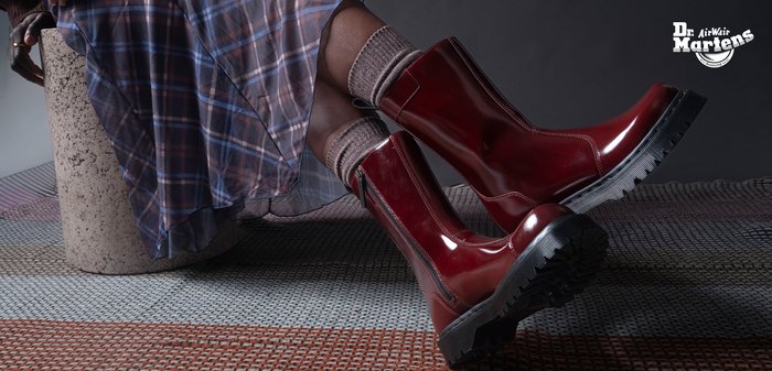 Persoon zittend met uitgestrekte benen, draagt glanzende bordeauxrode Dr. Martens-laarzen, beige sokken en een geruite rok, tegen een donkere achtergrond.