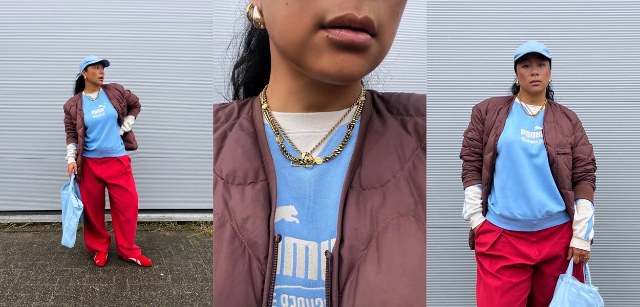 Vrouw met blauwe Puma-trui, rode broek, bruine jas, blauwe pet, gouden kettingen en een lichtblauwe totebag tegen een grijze muur.