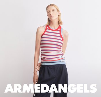 Donna con capelli biondi corti che indossa una canotta senza maniche a righe rosse, bianche e blu e pantaloni larghi scuri, su uno sfondo chiaro e uniforme.