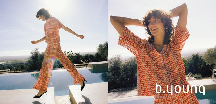 Lächelnde Frau mit lockigem Haar, die ein gemustertes orange-lila Kleid trägt und im Freien in der Nähe eines Pools mit Grünpflanzen im Hintergrund steht.
