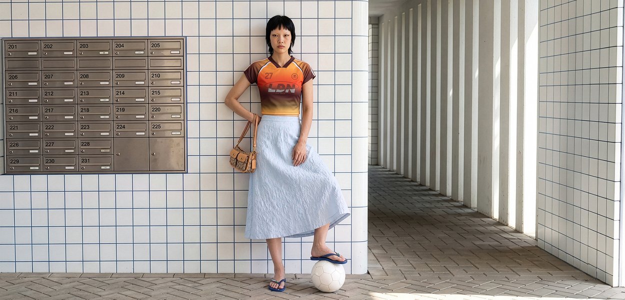 Mujer con camiseta deportiva naranja y falda azul claro de pie junto a una pared con azulejos, con el pie sobre un balón de fútbol blanco cerca de los buzones.