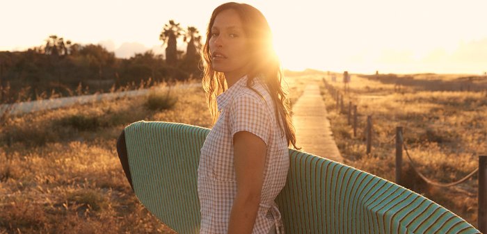 Vrouw die een gestreept surfplank draagt, wandelt op een houten pad bij zonsondergang door een graslandschap.