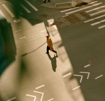 Een persoon in een lange, gele jas loopt op een straat met asfalt en werpt een schaduw; witte markeringen en schaduwen van omliggende gebouwen zijn zichtbaar.