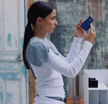 Langärmliges weißes Top mit grauen Stoffakzenten und weicher Textur. Die Person hält einen blauen Gegenstand und betrachtet ein weißes Modellgebäude.