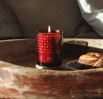 Bougie en verre rouge avec un motif doré métallique, flamme allumée et texte à l'avant. Placée sur un plateau en bois avec des pâtisseries à proximité.