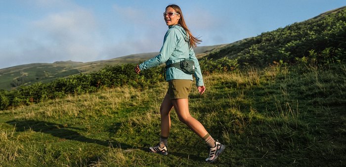 Jeune femme portant des lunettes de soleil et une veste, randonnée sur une colline herbeuse sous un ciel clair.