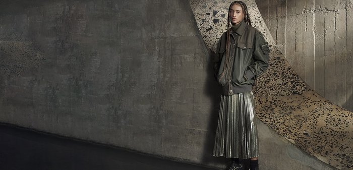 Giacca bomber verde con grandi tasche, abbinata a una gonna midi plissettata metallica. La texture morbida e gli stili contrastanti creano un interesse visivo.