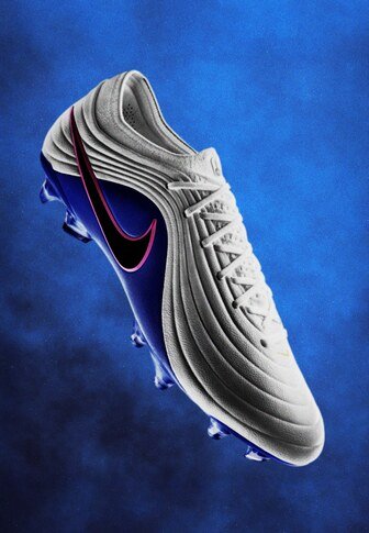 Witte en blauwe Nike voetbalschoen met roze swoosh, gestructureerde bovenzijde en veterluiting, tegen een blauwe verloopachtergrond.