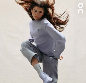 Hellblaue Sweatshirt mit Logo, kombiniert mit grauen Jogginghosen und weißen Schuhen. Das Model mit langen, fließenden Haaren posiert dynamisch.