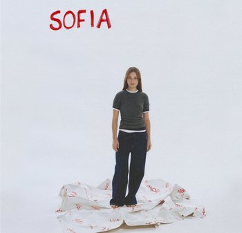 Mädchen trägt einen grauen kurzärmligen Pullover über einem weißen T-Shirt und blauen Jeans, steht auf einem zerknitterten weißen Textil mit rotem Text.