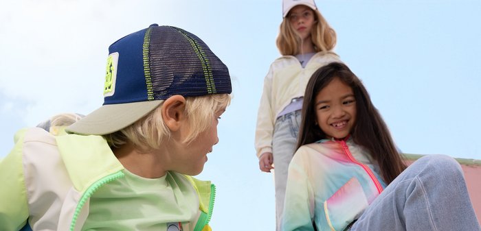 Tre bambini che indossano abiti casual colorati e cappellini, sorridenti e che interagiscono all'aperto con uno sfondo di cielo limpido.