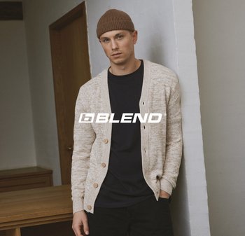 Hellbeige strukturierter Cardigan mit Holzknöpfen über einem schwarzen Hemd, getragen von einer Person vor einer neutralen Wand. Braune gestrickte Mütze auf dem Kopf.