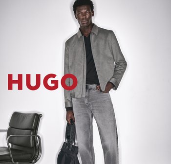 Mann in grauem Jackett und Jeans, der eine schwarze Tasche hält, steht neben einem schwarzen Stuhl mit rotem HUGO-Text auf weißem Hintergrund.