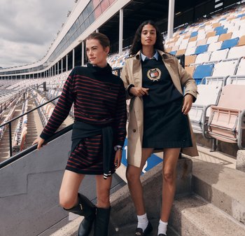 Zwei Frauen stehen auf den Stadionstufen. Eine trägt ein schwarz-rotes gestreiftes Sweaterkleid; die andere trägt einen blauen Pullover mit einem Emblem und einen beigen Trenchcoat.