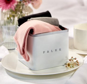 Weißes Metallbehälter mit dem Aufdruck "FALKE", gefüllt mit zusammengefalteten Socken in Rosa, Grau und Schwarz. Auf einem weißen Teller mit zarten Blumen platziert.