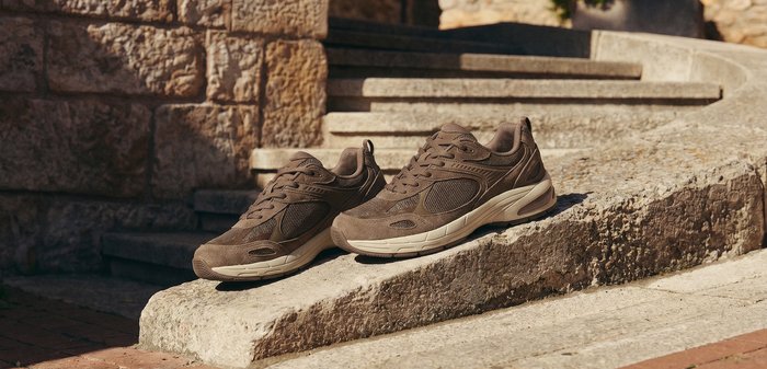 Pair of brown athletic sneakers placed on a stone ramp beside stone steps with a textured stone wall and potted plant in the background.