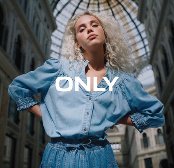 Jonge vrouw met krullend blond haar, gekleed in een blauw denim outfit, staat zelfverzekerd in een stedelijke arcade met glazen dak, met de tekst "ONLY" over de afbeelding.