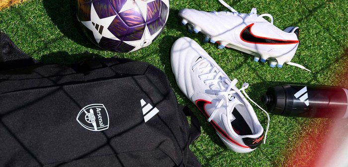 Valged Nike jalgpallijalatsid, lilla ja valge jalgpall, must Arsenal seljakott ja must Adidas joogipudel rohelisel murul.