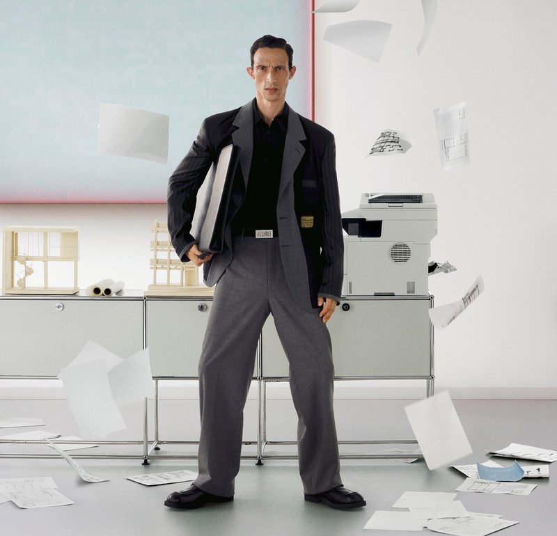 Hombre con traje gris sosteniendo una carpeta, de pie en una oficina con papeles volando y esparcidos alrededor de una fotocopiadora y armarios.