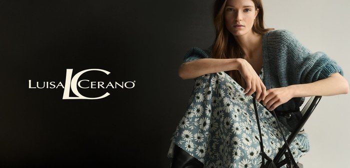 Knit teal cardigan over a floral-patterned dress with blue and white daisies. Model poses seated on a black chair against a dark backdrop.