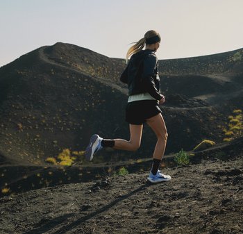 Femme avec une queue de cheval portant une tenue de sport noire courant en montée sur un terrain volcanique dans un environnement de montagne en plein jour.