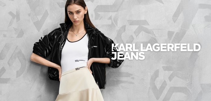 Femeie cu păr lung și castaniu, purtând un maiou alb cu logo, fustă crem și o jachetă neagră lucioasă, stând în fața unui perete gri texturat.