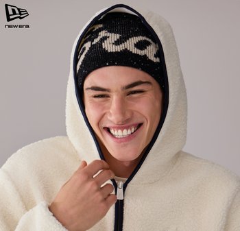 Hvid fleece-lynlåshættejakke med marineblå kant, kombineret med en sort strikhue med cream-mønster, båret af en smilende person.