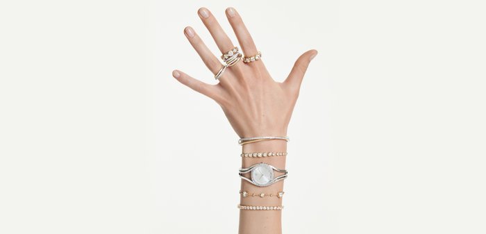 Hand met gouden en zilveren ringen en meerdere armbanden, waaronder een zilveren horloge, tegen een witte achtergrond.