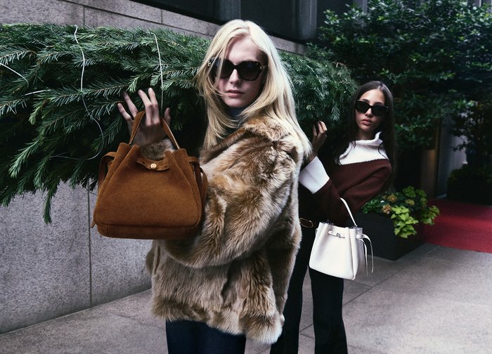 Brun mockaväska med rundad form och korta handtag. Modellen bär en fuskpälsjacka. Bakgrunden visar grönska och stadens arkitektur.