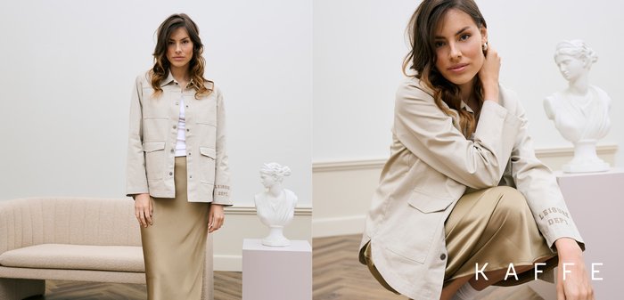 Mlajša ženska v bež jakni in krilu sedi v notranjosti, v ozadju pa je klasičen beli kip glave na podstavku.