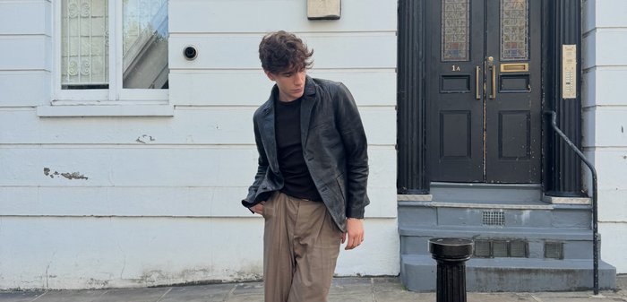 Giacca di pelle nera sopra una camicia nera, pantaloni beige a pieghe, in piedi su un marciapiede vicino a una parete bianca texturizzata e a una porta nera.