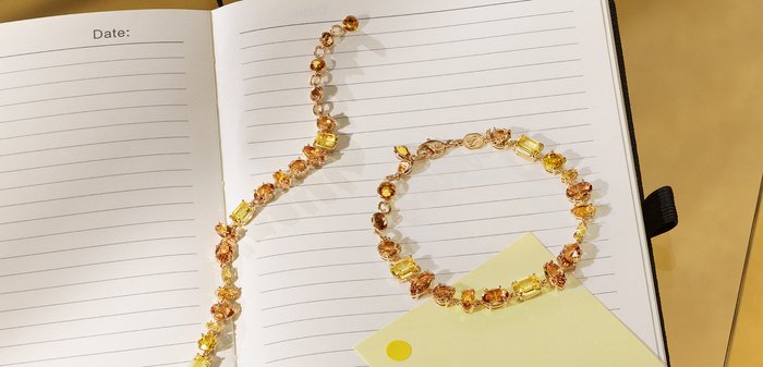 Goldfarbener Armreif und passendes Halsband mit mehreren bernsteinfarbenen und gelben Edelsteinen in ovalen und rechteckigen Formen, die auf einem Notizbuch liegen.