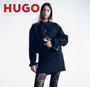 Joven mujer con chaqueta negra de gran tamaño, minifalda, medias transparentes con estampado floral, sosteniendo un bolso de mano negro, de pie frente a un fondo liso.