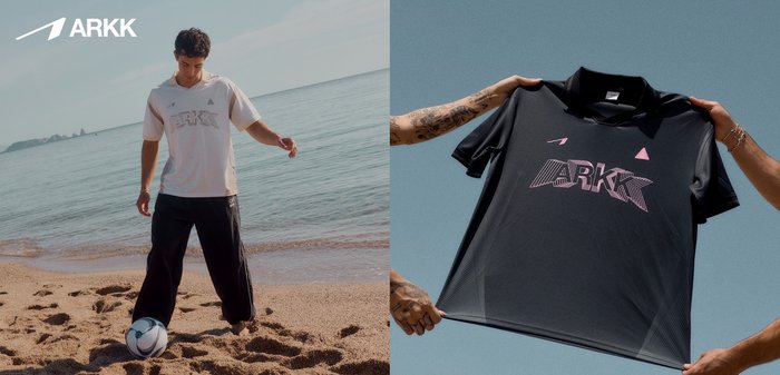 Ung mand iført ARKK-shirt, der leger med en fodbold barfodet på en sandstrand ved det rolige hav under en klar himmel.