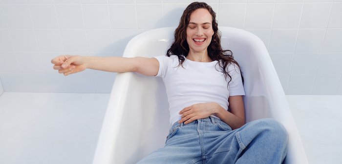 Frau mit lockigem Haar, gekleidet in weißes Hemd und Jeans, entspannt sich mit geschlossenen Augen und lächelt, während sie in einer leeren weißen Badewanne sitzt.