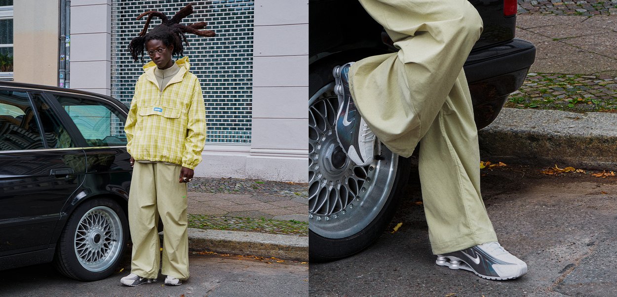 Persoană cu dreadlocks, purtând ochelari, un sacou cu carouri galbene, pantaloni bej și adidași Nike, stând lângă o mașină neagră pe trotuar.
