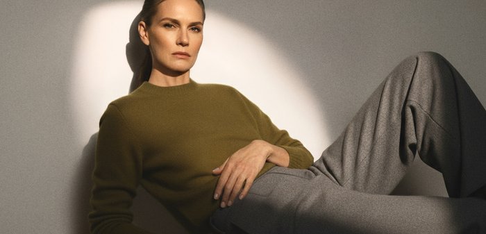 Sieviete atpūšas, atslābinoties pret sienu, valkājot olīvu zaļu džemperi un pelēkas plaša piegriezuma bikses mīkstā apgaismojumā.