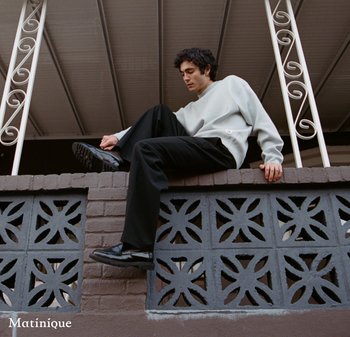 Young man in white sweater and black pants sitting on a decorative brick wall under a covered porch, adjusting his shoe.