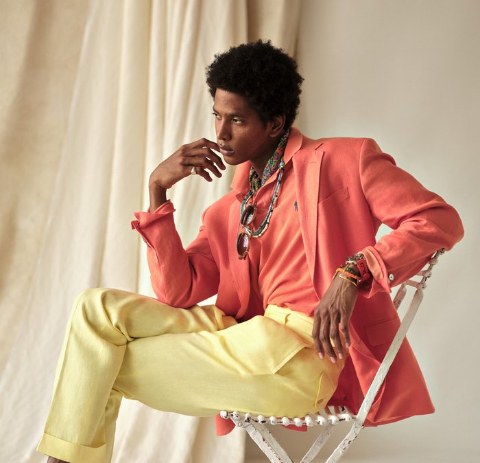Mann sitzt auf weißem Stuhl, trägt einen korallenfarbenen Blazer, orangefarbenes Hemd, gelbe Hose, Sonnenbrille, Halskette und mehrere Armbänder vor beigefarbenem Hintergrund.