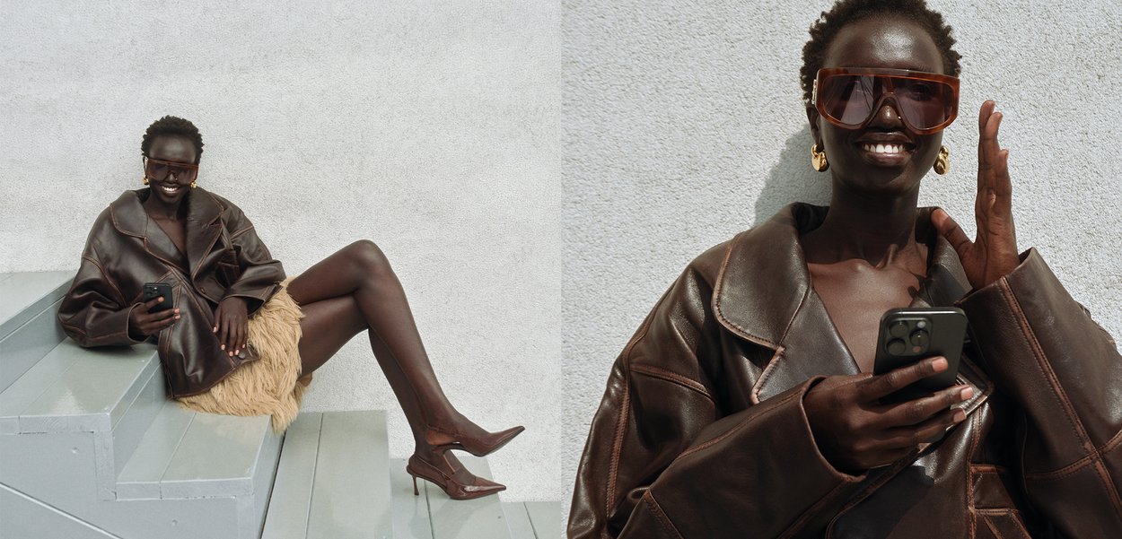 Brown leather oversized jacket, textured tan mini skirt, transparent oversized sunglasses, and pointed brown heels. Sitting on grey stairs.